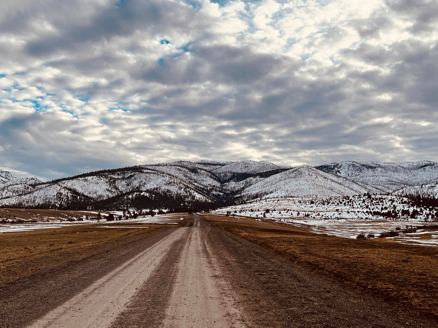 Picture of mountains in the distance with clouds, snow and a road.